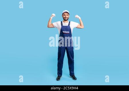 Full length cheerful handyman in overalls, cap and protective gloves standing, showing biceps, demonstrating strength. Profession of service industry, Stock Photo