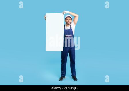 Full length of cheerful labourer man in workwear and protective gloves raising vertical sheet poster template, mock up blank space for advertising mes Stock Photo