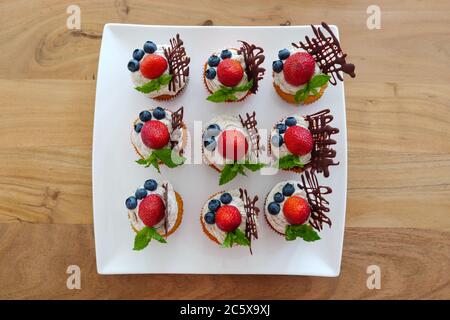 Top view at colourful group of cupcakes decorated with white whip cream, strawberries, blueberries, chocolate and mint on white plate and wood table. Stock Photo