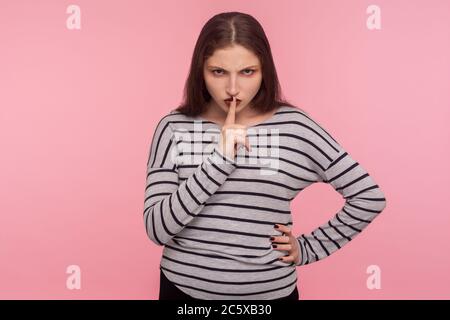 Shut up! Portrait of angry displeased woman in striped sweatshirt saying hush, be quiet with finger on lips gesture, keep silence, don't tell secret. Stock Photo