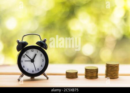Alarm clock and stacks of coins on wooden table, natural green blurred background. Business, finance, time, online shopping, money saving concept. Stock Photo