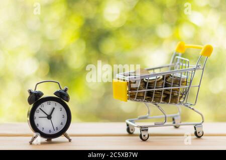 Alarm clock and mini shopping cart full of coins on wooden table, natural green blurred background. Business, finance, time, online shopping, money sa Stock Photo