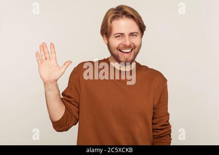 Portrait of hospitable friendly bearded man in sweatshirt raising hand waving hello hi and smiling, happy to meet friend, welcoming or gesturing goodb Stock Photo