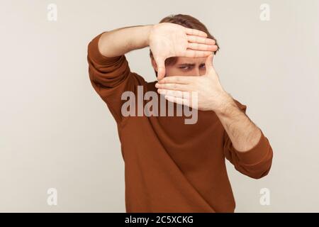 Portrait of creative man in casual sweatshirt looking through hand frame gesture, cropping image, zooming and focusing on distance, viewing target. in Stock Photo