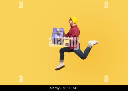 Enthusiastic extremely joyful hipster guy running quickly fast with gift box, flying in air holding birthday present surprise, hurry to give holiday b Stock Photo