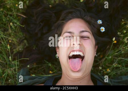 Portrait of a young woman lying in the grass with flowers in her hair. She sticks out her tongue with a happy expression Stock Photo