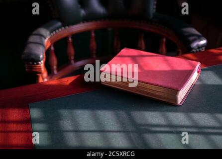 Closed bible on the edge of table with chesterfield chair at the background. Light play forming lines and patterns on surface. Stock Photo