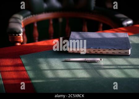 Closed bible on the edge of table with chesterfield chair at the background. Light play forming lines and patterns on surface. Stock Photo