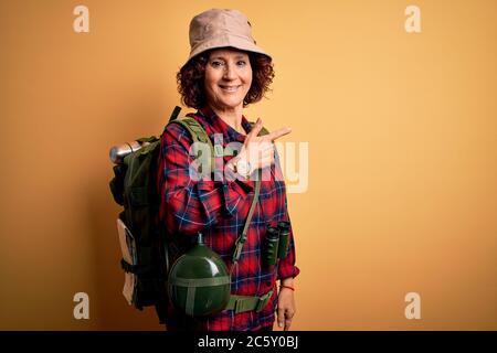 Middle age curly hair hiker woman hiking wearing backpack and water canteen using binoculars cheerful with a smile on face pointing with hand and fing Stock Photo