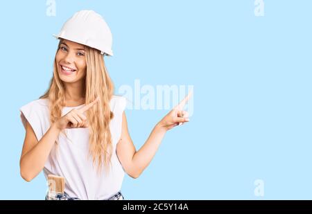 Beautiful caucasian woman with blonde hair wearing hardhat and painter clothes smiling and looking at the camera pointing with two hands and fingers t Stock Photo