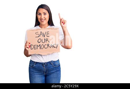 Young beautiful woman holding save our democracy cardboard banner surprised with an idea or question pointing finger with happy face, number one Stock Photo
