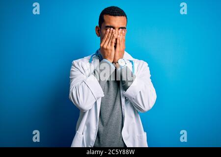 Handsome african american doctor man wearing coat and stethoscope over blue background rubbing eyes for fatigue and headache, sleepy and tired express Stock Photo