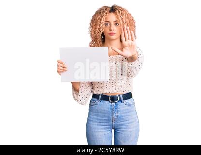 Young blonde woman with curly hair holding sale poster with open hand doing stop sign with serious and confident expression, defense gesture Stock Photo