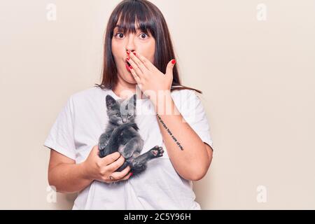 Young plus size woman holding cat covering mouth with hand, shocked and afraid for mistake. surprised expression Stock Photo