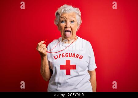 Senior beautiful grey-haired lifeguard woman wearing t-shirt with red cross using whistle sticking tongue out happy with funny expression. Emotion con Stock Photo