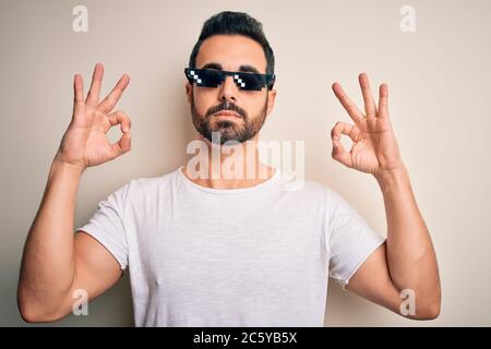 Young handsome man with beard wearing funny thug life sunglasses over white background relax and smiling with eyes closed doing meditation gesture wit Stock Photo
