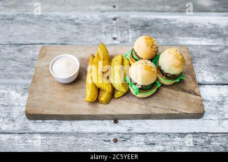 Three mini burgers with beef patty, french fries and mayonnaise on wooden board. Shabby aged table background. Unusual food serving. Fast food and unhealthy lifestyle. Stock Photo