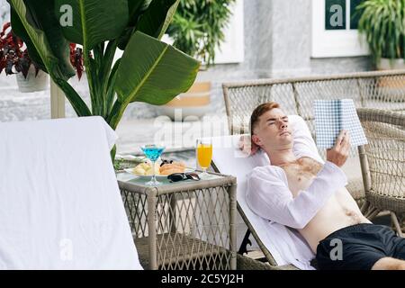 Handsome young man resting on chaise-lounge, eating breakfast and reading interesting book Stock Photo