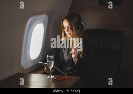 Attractive businesswoman using digital tablet in private plane. Lovely caucasian young woman in stylish black dress listening music with wireless head Stock Photo