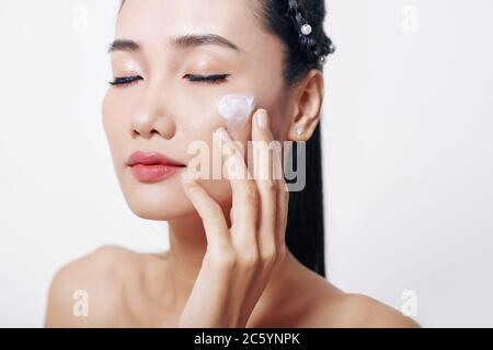 Young Asian woman closing eyes and applying anti-aging mosturizing soft cream o her face Stock Photo