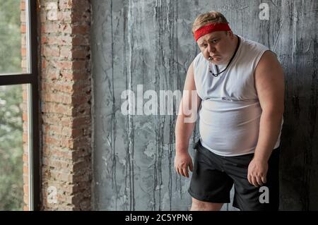 tired fat caucasian man after sport exercises, man is exhausted after fitness, he holds a whistle in his mouth and looks at camera with sadness Stock Photo