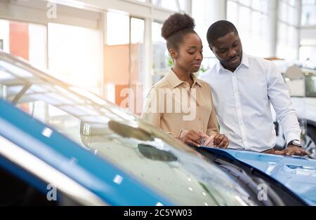 confident handsome afro man shows to wife a car he is liked, they look at car and discuss, man and woman think before making purchase Stock Photo