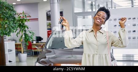 portrait of young and beautiful black woman with keys from her new car, she is happy in dealership, luxurious car in the background Stock Photo