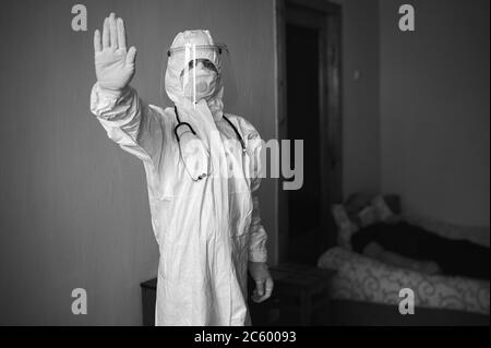 Black and white image of doctor in PPE suit, face mask, gloves, phonendoscope, visiting patient at home during covid-19 quarantine, showing stop sign, lying patient on background Stock Photo