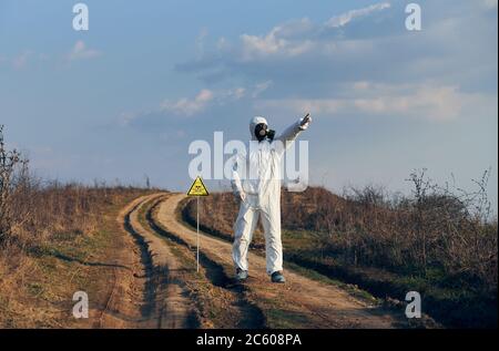 Environmentalist standing on the road near yellow triangle with skull and crossbones warning sign and pointing finger at sky. Research scientist wearing protective suit, gas mask and shoe covers. Stock Photo