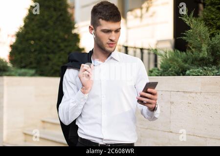 Young handsome man in white shirt with wireless earphones holding black jacket on shoulder while thoughtfully using cellphone outdoor Stock Photo