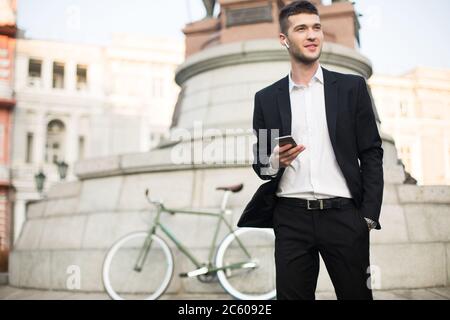 Young handsome businessman in classic black suit and white shirt with wireless earphones holding cellphone in hand while dreamily looking aside with Stock Photo