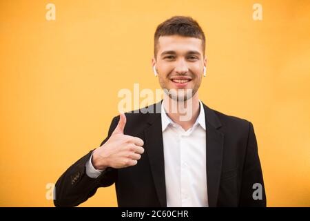 Young smiling man in classic black jacket and white shirt with wireless earphones happily showing thumb up gesture while looking in camera over orange Stock Photo