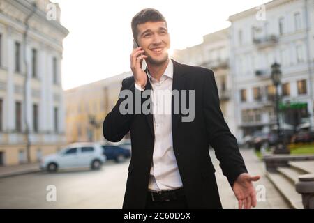 Young joyful businessman in black jacket and white shirt happily talking on cellphone while looking aside with beautiful city view on background Stock Photo