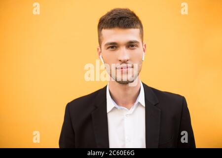 Portrait of young handsome man in black jacket and white shirt with wireless earphones thoughtfully looking in camera over orange background Stock Photo