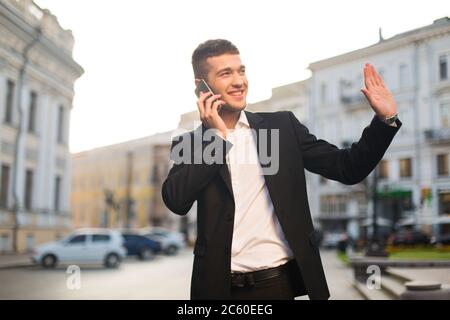 Young handsome man in black jacket and white shirt happily looking aside and waving while talking on cellphone with beautiful city view on background Stock Photo