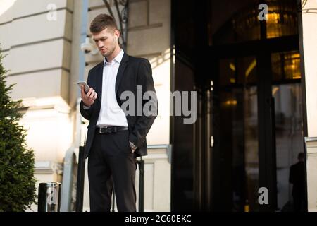 Young serious businessman in black suit and shirt with wireless earphones thoughtfully using cellphone outdoor Stock Photo