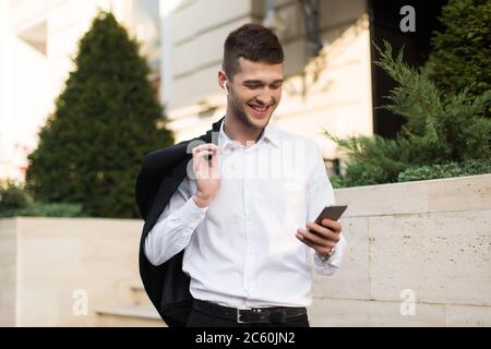 Young cheerful man in white shirt with wireless earphones holding black jacket on shoulder while happily using cellphone outdoor Stock Photo