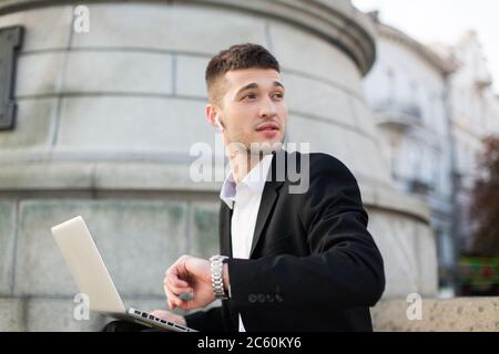 Young pensive businessman in black suit with wireless earphones dreamily looking aside with hand watch and laptop on knees outdoor Stock Photo