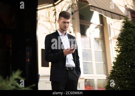 Young thoughtful businessman in black suit and shirt with wireless earphones holding cellphone in hand with beautiful building on background Stock Photo