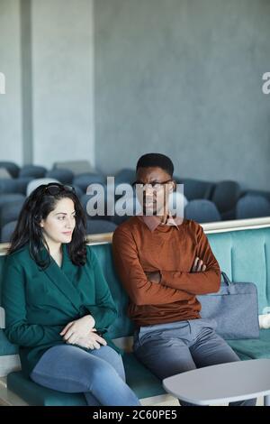 Vertical portrait of young African-American man sitting with Middle-Eastern woman sitting on chairs in auditorium while waiting for seminar or class, Stock Photo