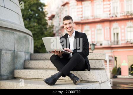 Young smiling businessman in classic black suit with wireless earphones joyfully looking in camera working with laptop on little stairs while spending Stock Photo