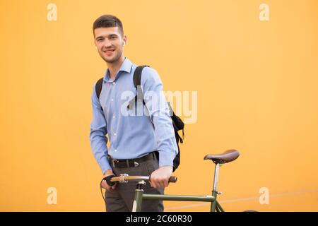 Young attractive man in blue shirt and backpack with wireless earphones standing with retro bicycle while dreamily looking in camera over orange Stock Photo