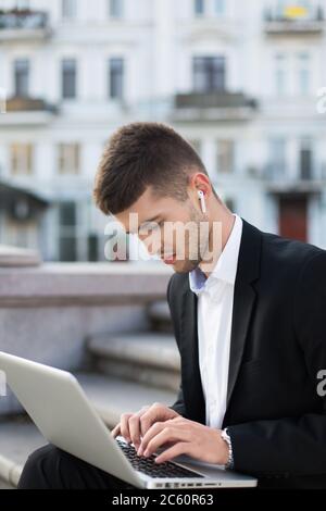 Young pensive businessman in classic black suit with wireless earphones thoughtfully working on laptop while spending time outdoor Stock Photo