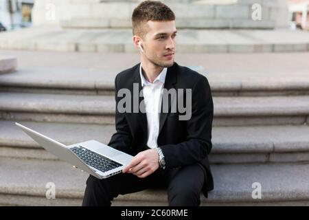 Young pensive businessman in classic black suit with wireless earphones thoughtfully looking aside holding laptop in hands while spending time outdoor Stock Photo