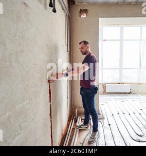 Young man in empty unfinished apartment measuring the wall with a help of measure tape, standing on floor heating pipe system against panoramic window. Concept of refurbishment and renovation. Stock Photo