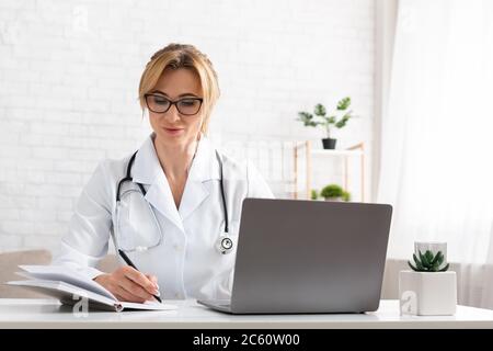 Family doctor online. Busy woman writes in notebook, near laptop Stock Photo
