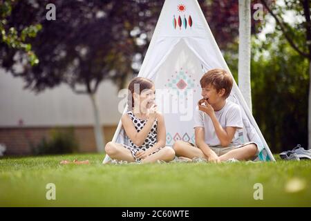 Cute happy little children sitting in teepee at backyard and eats cookies. Stock Photo