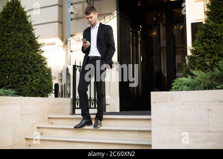 Young attractive businessman in black suit and white shirt with wireless earphones thoughtfully using cellphone while going down the stairs with Stock Photo