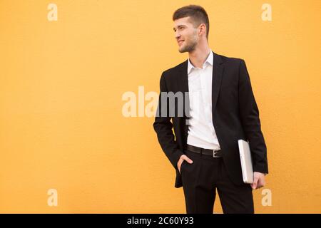 Young smiling man in classic black suit and white shirt holding laptop in hand while dreamily looking aside over orange background Stock Photo