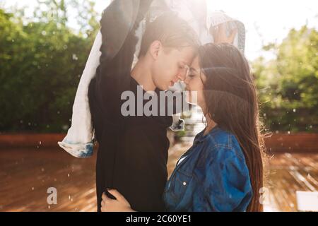 Portrait of young smiling couple covering themselves with denim jacket while standing under rain in park Stock Photo